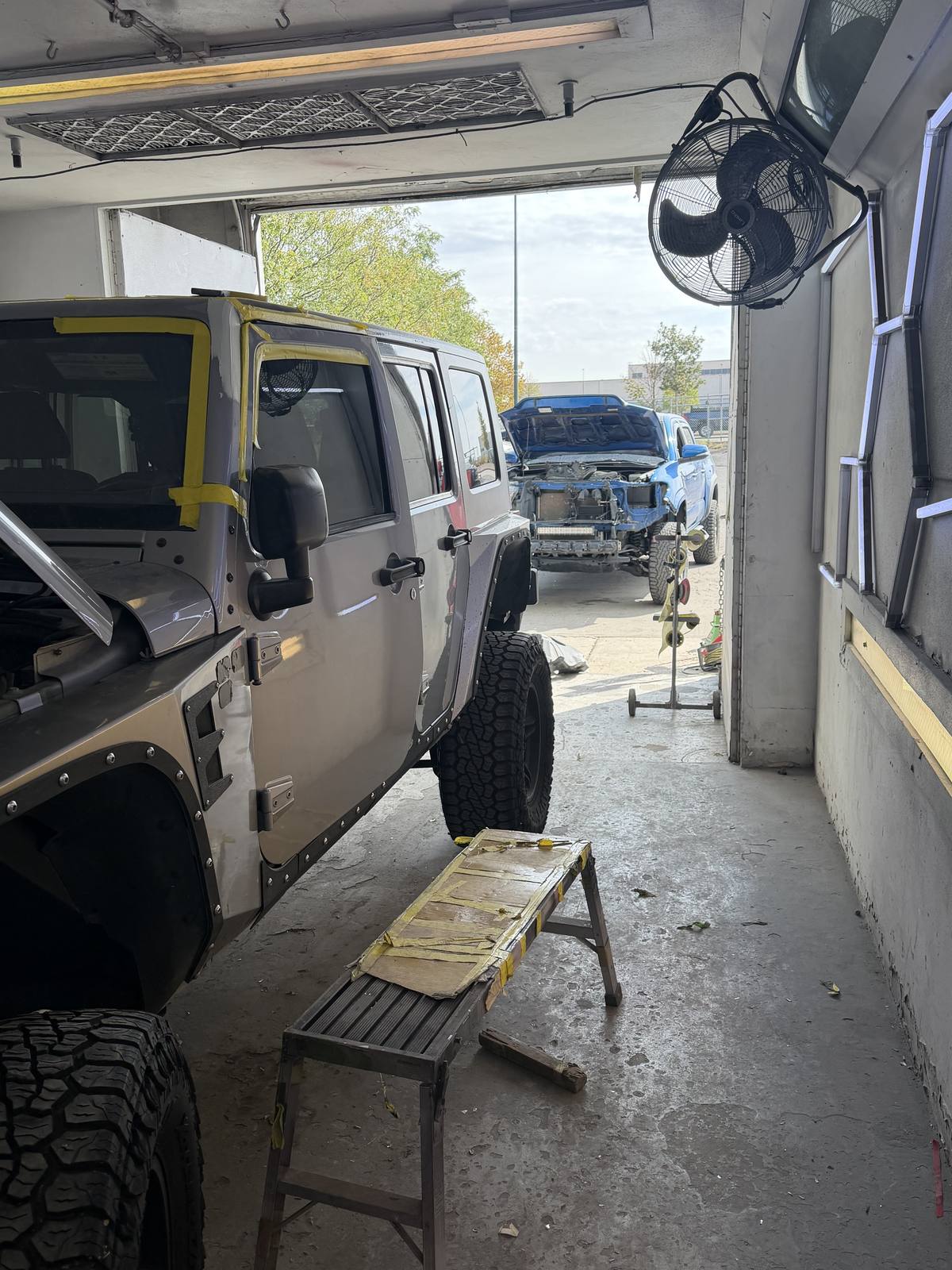 A Jeep Wrangler masked with tape in the bay, and a pickup with its front end stripped on the lot outside.