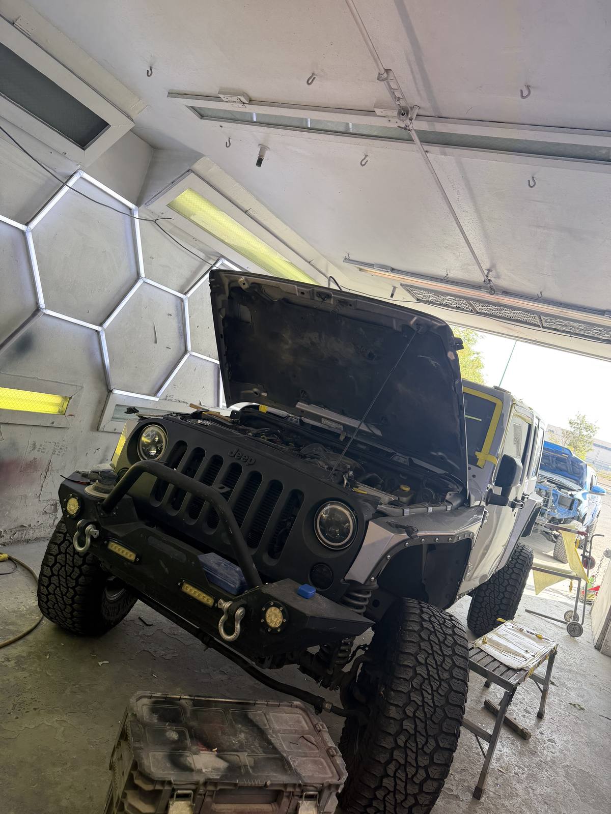 A Jeep Wrangler in the bay with the hood raised, under the shop's overhead lighting.