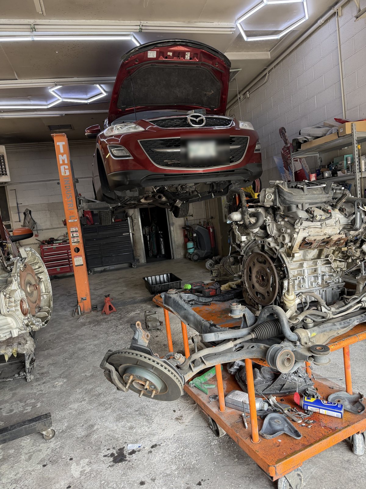 A Mazda on the lift at Auto Tek with its engine and subframe on a cart during mechanical repair.
