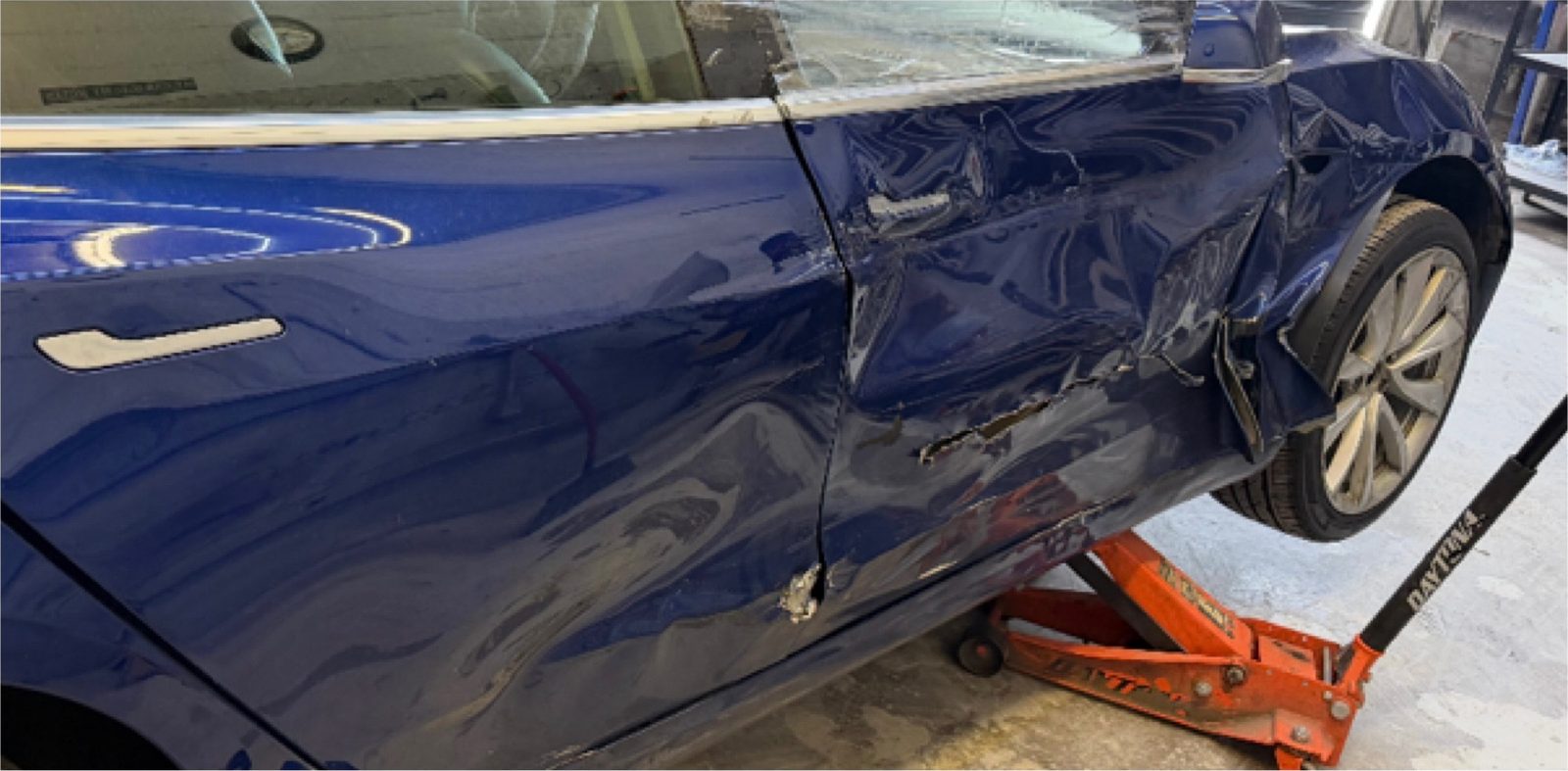 Blue Tesla Model 3 with heavy impact damage to the rear door and quarter panel, raised on a jack in the bay.