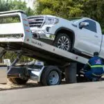 Tow truck operator loading white GMC pickup truck on street in daytime.