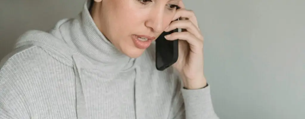 Concentrated young female in gray domestic clothes browsing netbook and having conversation via cellphone in light room