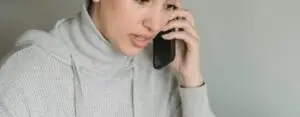 Concentrated young female in gray domestic clothes browsing netbook and having conversation via cellphone in light room