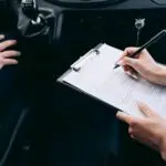 Close-up of a person writing on a clipboard inside a car, showing hands and a gear shift.